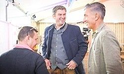Drei Männer stehen in einem Raum. Der Mann in der Mitte lacht, während der Mann auf seiner linken Seite lächelt und der Mann auf seiner rechten Seite zusieht.