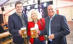 Drei Personen posieren für ein Foto bei einem Bierfest. Zwei Männer und eine Frau, alle formell gekleidet, halten Bierkrüge hoch. Der Hintergrund zeigt eine Bühne mit Lautsprechern und einem Banner.