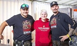 Drei Personen stehen in einem Raum mit weißen Wänden. Zwei Männer tragen schwarze T-Shirts, während die Frau in der Mitte eine rote Kapuzenjacke trägt. Die Männer haben Tattoos an den Armen.