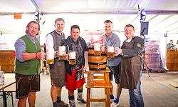 Fünf Männer in traditioneller bayerischer Kleidung stehen um ein großes hölzerne Fass und halten Bierkruge hoch. Sie befinden sich wahrscheinlich auf einem Bierfestival oder einer traditionellen deutschen Veranstaltung.