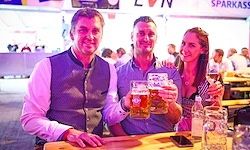 Drei Personen posieren mit Bierkrügen in der Hand an einer Bar. Ein Mann trägt eine Weste, ein anderer hält ein Telefon und eine Frau ein Glas. Im Hintergrund sind weitere Gäste und ein Veranstaltungsbanner zu sehen.