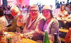Ein Mann und eine Frau sitzen zusammen an einem überfüllten Tisch, lächeln und posieren für ein Foto. Der Mann hält ein großes Glas Bier, und sie sind von verschiedenen Flaschen und Gläsern umgeben.