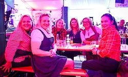 Eine Gruppe von Frauen in traditioneller bayerischer Kleidung sitzt an einem Tisch in einer Bar, lächelt und posiert für ein Foto.