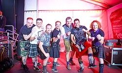 Sieben Männer in Kilts posieren für ein Foto auf einer Bühne mit roter Beleuchtung im Hintergrund.