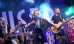 Eine Band spielt auf der Bühne, ein Mitglied spielt Akkordeon, ein anderes Gitarre und ein drittes Schlagzeug. Die Bühne ist mit verschiedenen Instrumenten und einem Hintergrund ausgestattet.