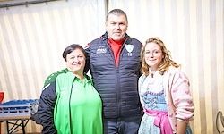 Drei Menschen posieren für ein Foto in einem Innenbereich. Zwei Frauen und ein Mann stehen nebeneinander. Der Mann trägt ein rotes Shirt und eine Jacke. Eine Frau trägt ein grünes Kleid und die andere ein rosa Kleid.