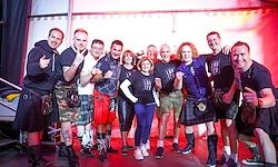 Eine Gruppe von Menschen in Kilts und T-Shirts posiert auf einer Bühne mit roter Beleuchtung. Einige tragen Leder und Sneakers, und sie alle scheinen zu lächeln.