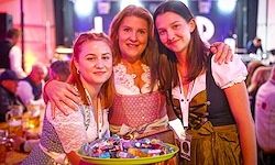 Drei Frauen stehen lächelnd bei einer Veranstaltung zusammen. Die Frau auf der linken Seite hält einen grünen Teller mit Süßigkeiten und die mittlere Frau hat ihren Arm um die Schultern der beiden anderen gelegt. Im Hintergrund ist eine verschwommene Menge mit Tischen und Stühlen zu sehen.