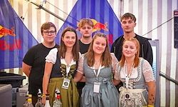 Eine Gruppe von Menschen in traditioneller deutscher Kleidung posiert für ein Foto vor einem Hintergrund mit Fahnen und Treppen. Sie haben Namensschilder und scheinen auf einem Festival zu sein.