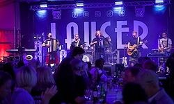 Eine Band tritt auf einer Bühne mit lila Beleuchtung auf. Das Wort 'Lauser' ist prominent auf einem Banner hinter der Band zu sehen. Die Bandmitglieder spielen verschiedene Instrumente, darunter Gitarren und Akkordeons. Das Publikum sitzt vor der Bühne und beobachtet die Aufführung.