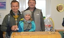 Drei Personen stehen in der Nähe einer Holzkiste; der Mann auf der linken Seite hält eine Trophäe, der Mann in der Mitte trägt eine Weste und ein Mädchen in einer Mütze steht zwischen ihnen. Sie befinden sich in einem Raum mit einer weißen Wand und einem runden Objekt über ihnen.