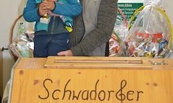 Eine Holzkiste mit dem Wort 'Schwadorfer' darauf, dahinter stehen zwei Personen und im Hintergrund befinden sich verschiedene Plastiktüten mit Gegenständen darin.