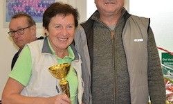 Zwei Erwachsene stehen zusammen und lächeln. Die Frau hält eine goldene Trophäe. Sie sind drinnen, mit gerahmten Bildern an der Wand hinter ihnen.