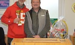 Zwei Männer stehen vor einem Schreibtisch mit Trophäen und einem Geschenkbeutel. Der Mann auf der linken Seite hält einen Siegerpokal. Der Mann auf der rechten Seite lächelt. Dahinter ist eine weiße Wand mit einer kreisförmigen Dekoration.