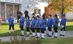 Eine Marschkapelle in blauen Uniformen und Hüten steht vor einem Gebäude, wobei Zuschauer im Hintergrund zu sehen sind.