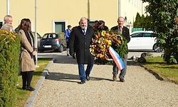 Zwei Männer tragen einen Kranz mit einem dreifarbigen Band und gehen vor einem Gebäude mit Menschen und geparkten Autos her.