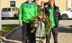 Zwei Männer in grünen Jacken tragen einen Kranz. Sie stehen vor geparkten Autos und einem Gebäude.