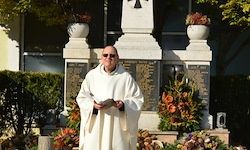 Ein Priester steht vor einem Denkmal, ein Buch in der Hand, in einer Sonnenbrille und einem weißen Gewand.