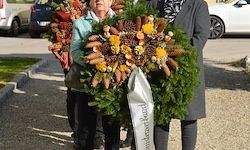 Drei Personen tragen Kränze, einer mit Tannenzapfen und Blumen, einen Pfad entlang, mit geparkten Autos im Hintergrund.