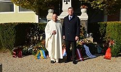 Zwei Männer stehen vor einem Gebäude, einer in einem weißen Gewand und der andere in einem schwarzen Mantel. Sie sind von Blumen und Flaggen umgeben, mit einer Statue hinter ihnen.