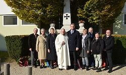 Eine Gruppe von Menschen, darunter ein Priester, steht vor einem Denkmal mit einem Kreuz und Büschen im Hintergrund.