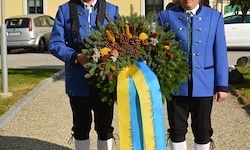 Zwei Männer in blauen Uniformen stehen vor einem Gebäude mit geparkten Autos und halten einen Kranz mit einem gelben und blauen Band.