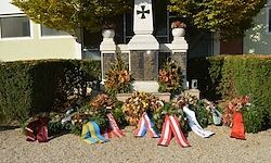 Ein Denkmal mit einem Kreuz auf der Spitze, umgeben von verschiedenen farbigen Bändern und Blumen, vor einem Gebäude mit Bäumen platziert.