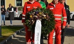 Zwei Personen in roten Sicherheitsanzügen halten einen Kranz, während mehrere andere in Freizeitkleidung im Hintergrund stehen, in der Nähe einiger geparkter Autos.
