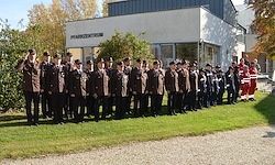 Eine Reihe uniformierter Personen steht vor einem Gebäude, einige tragen Hüte und Schuhe, andere Stiefel. Dahinter ist eine Person in einem rot-weißen Outfit zu sehen. Im Hintergrund befinden sich Bäume und Pflanzen.