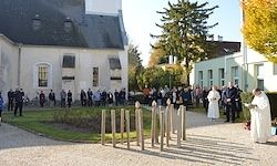 Eine Menschenmenge versammelt sich vor einer Kirche, wobei Geistliche in weißen Roben neben einer hölzernen Barriere stehen. Die Umgebung umfasst Herbstbäume und einen klaren Himmel.