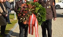 Zwei uniformierte Personen tragen einen Kranz mit roten und weißen Bändern vor einer Gruppe von Menschen und einem geparkten Auto.