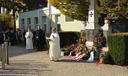 Ein Priester in Weiß steht vor einem Gedenkort mit Blumen und Kränzen, vor dem mehrere Menschen beobachten. Hinter ihm steht ein Gebäude mit einem Kreuz an der Wand.