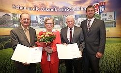 Vier Personen stehen vor einem grünen Hintergrund mit einem Banner. Sie halten Urkunden. Die Frau in der Mitte hält einen Blumenstrauß. Das Banner lautet Schau in die Zukunft, sich Schwador.