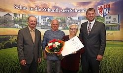 Drei Menschen stehen vor einem Poster mit Text, halten einen Blumenstrauß und ein Zertifikat in der Hand. Sie lächeln und posieren für ein Foto.