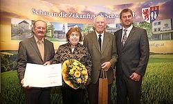 Eine Frau mit Blumen und einem Zertifikat steht zwischen zwei Männern, alle formal gekleidet vor einem grünen Feld. Dahinter ist ein Banner mit Text und Logos.