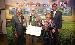 Eine Gruppe von vier Personen steht vor einem grünen Feld. Ein Mann hält ein Zertifikat und Blumen, während eine Frau Blumen hält. Sie alle scheinen zu feiern.