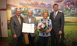 Vier Personen stehen vor einem Banner mit einem deutschen Logo. Der Mann und die Frau auf der linken Seite halten Blumen und ein Zertifikat. Die beiden Männer auf der rechten Seite tragen Anzüge und Krawatten.