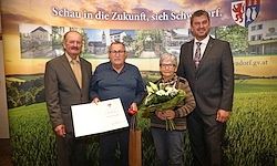 Drei Personen stehen vor einer Kulisse einer ländlichen Landschaft. Die mittlere Figur hält einen Blumenstrauß und ein Zertifikat. Der Mann auf der linken Seite trägt einen Anzug und hält ein weiteres Zertifikat. Der Mann auf der rechten Seite trägt einen Anzug und eine Krawatte.