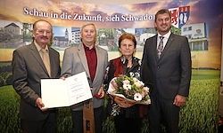 Vier Personen in formeller Kleidung stehen nebeneinander, halten ein Zertifikat und einen Blumenstrauß. Sie stehen vor einer Kulisse mit einem grünen Feld und Gebäuden.