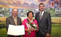 Drei Menschen stehen eng beieinander und lächeln. Ein Mann hält ein Papier, und eine Frau hält einen Blumenstrauß. Hinter ihnen wird ein Banner mit Text an einer Wand angezeigt.