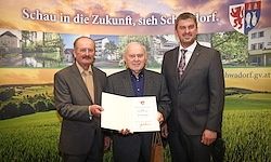 Drei Männer in Anzügen stehen nebeneinander und halten eine Urkunde. Sie posieren für ein Foto vor einer Kulisse mit Banner und grünem Feld.