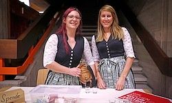 Zwei Frauen in traditioneller deutscher Kleidung stehen hinter einem Tisch mit verschiedenen Gegenständen, einschließlich einer Box mit der Aufschrift 'Spende'. Sie befinden sich in der Nähe einer Treppe mit Holzgeländern.