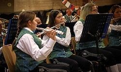 Eine Gruppe von Musikern spielt Instrumente in einem Raum mit Stühlen und Notenständern. Die Frau vorne spielt die Flöte.