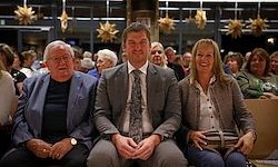 Drei Personen sitzen vor einer Menge in formeller Kleidung. Zwei Männer und eine Frau, der Mann auf der rechten Seite trägt eine Krawatte.