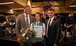 Drei Personen stehen auf einer Bühne mit einer Band im Hintergrund. Eine Frau hält ein Zertifikat und einen Blumenstrauß. Zwei Männer stehen zu ihrer Seite.