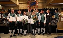 Eine Gruppe von Personen in traditioneller Kleidung steht auf einer Bühne mit Musikinstrumenten im Hintergrund. Sie halten Urkunden und Blumen und posieren für ein Foto.