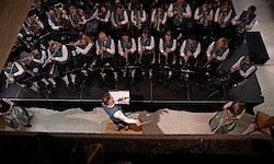 Ein großes Orchester spielt auf der Bühne mit einem Dirigenten, umgeben von Musikern in traditioneller Kleidung, die verschiedene Instrumente spielen. Eine Frau sitzt davor und schaut zu.