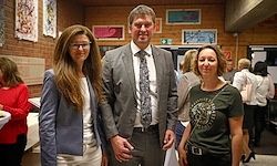 Drei Personen stehen in einem Innenraum zusammen, wahrscheinlich bei einer Veranstaltung. Ein Mann in formeller Kleidung steht zwischen zwei Frauen. Im Hintergrund ist eine mit verschiedenen Kunstwerken geschmückte Wand zu sehen, und Menschen unterhalten sich.