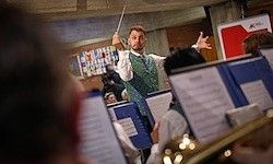Ein Mann dirigiert ein Orchester mit einem Taktstock, vor einer großen Gruppe von Musikern stehend. Das teilweise verschwommene Publikum sitzt vor ihm. Musikinstrumente sind sichtbar, und einige Dekorationen hängen an der Wand.