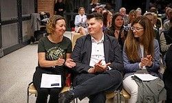 Drei Personen sitzen auf Stühlen und klatschen. Eine Frau links lächelt, ein Mann in der Mitte zeigt mit den Händen, und eine Frau rechts trägt eine Brille. Dahinter sitzen und stehen Personen, und eine Gebäudewand ist sichtbar.